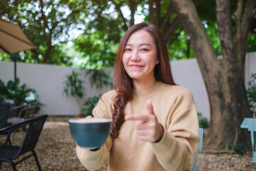 Portrait image of a woman holding and pointing finger at a cup of coffee in the outdoors cafe