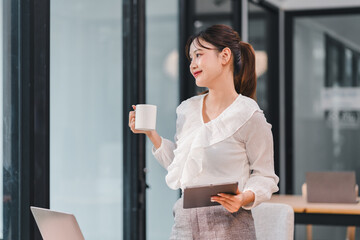 Young woman holding coffee cup and digital tablet in modern office with laptop on table, relaxed and thoughtful expression
