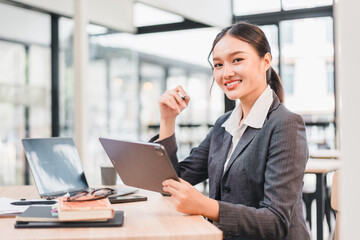 Fototapeta premium Young businesswoman holding digital tablet and smiling confidently in modern office with laptop and books on desk