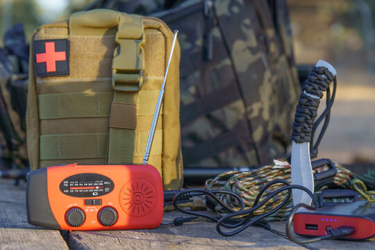 Survival kit including emergency crank radio, first aid kit, knife, paracord bracelet and power bank are lying on wooden table - Powered by Adobe