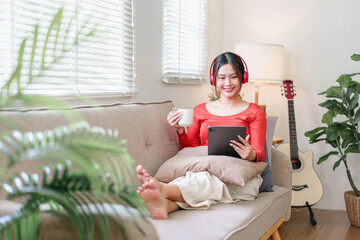 Young woman relaxing on sofa with tablet and coffee cup, wearing red headphones and smiling in cozy...