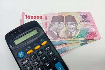 A dark-colored calculator with yellow and blue buttons is positioned next to a fan of Indonesian...