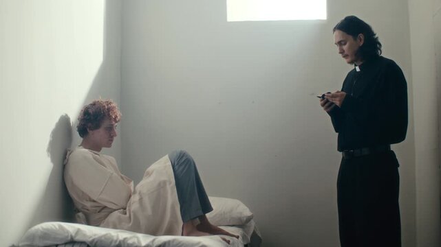 Young patient in straightjacket sitting on hospital bed, listening to priest reading scripture from Bible in sunlit psychiatric ward