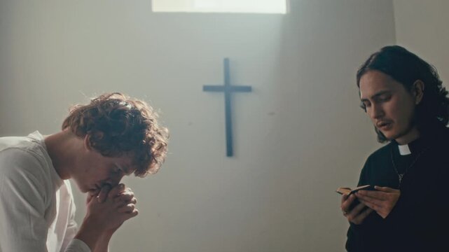 Young psychiatric patient clasping hands and bowing his head in prayer as priest reading scripture from book in sunlit mental hospital ward with cross on wall. Zoom shot