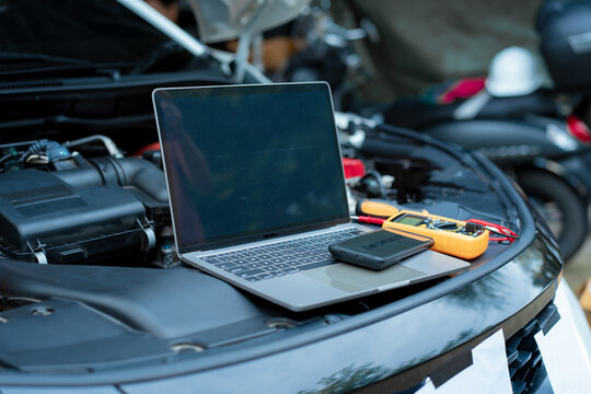 Essential car maintenance tools set up: a laptop rests on the open engine bay, a smartphone displaying “CAR CHECKLIST” on top, and a multimeter with test leads placed nearby