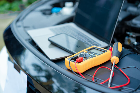 Essential car maintenance tools set up: a laptop rests on the open engine bay, a smartphone displaying “CAR CHECKLIST” on top, and a multimeter with test leads placed nearby