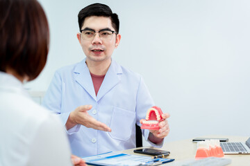 An Asian male dentist explains dental health to a middle-aged Asian female patient while holding a...