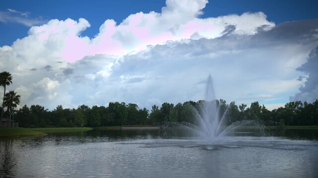 Fountain sprays high against a dramatic cloud-filled sky over a tranquil lake, filmed in 4K, 24fps