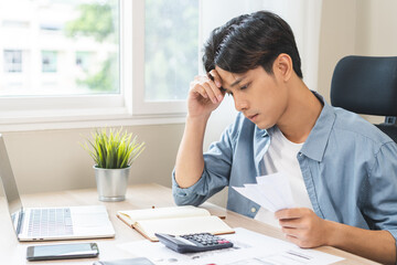 Debt and financial concept, stressed asian young man writing a plan finance while male sitting calculate expenses, income, hand holding bills for prepare payment or pay bills on table at home.