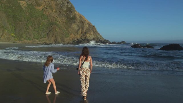 other and daughter standing hand in hand at the edge of the ocean, enjoying waves at Pfeiffer Beach in Big Sur, California.
