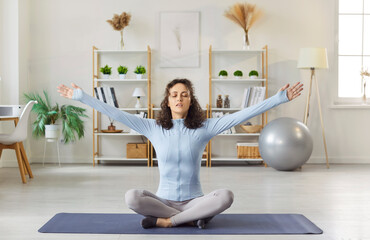 Indoor shot of healthy relaxed woman sitting crossed legs with arms raised on fitness mat,...