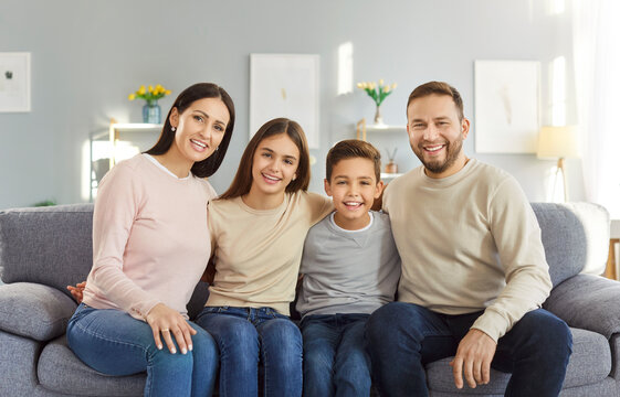 Portrait of happy family of four sitting together on sofa in living room, hugging and looking at camera. Smiling parents and kids embracing, posing for photo, showing closeness and happiness at home.