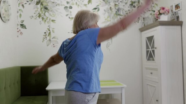 Senior woman wearing comfortable sportswear standing in living room, performing arm circles exercise targeting upper body mobility and enhancing flexibility for active aging health