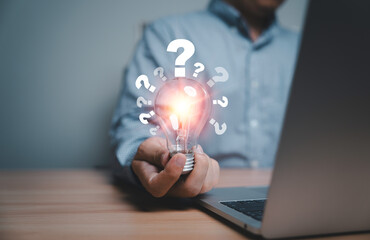Businessman using laptop and holding glowing lightbulb and question mark with copy space for...
