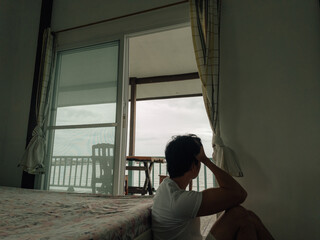 Thai man sitting near bed looking outside through window curtains with pensive emotion