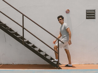 Confident Thai man in casual grey outfit walking beside outdoor steel staircase on sunny day