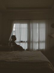 Thai man alone in a bedroom backlit by curtain light, expressing solitude and peace