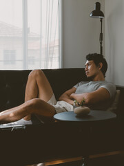 Thai man lying with legs stretched out on sofa in peaceful natural daylight setting in a quiet minimalist Thai apartment with natural daylight