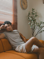 Young Thai man reclining on orange sofa looking thoughtful in cozy daylight room in a quiet minimalist Thai apartment with natural daylight