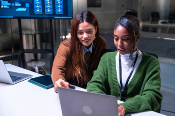Two programmer women working together in office environment focused on code development and software projects while using laptop and discussing ideas with enthusiasm and collaboration