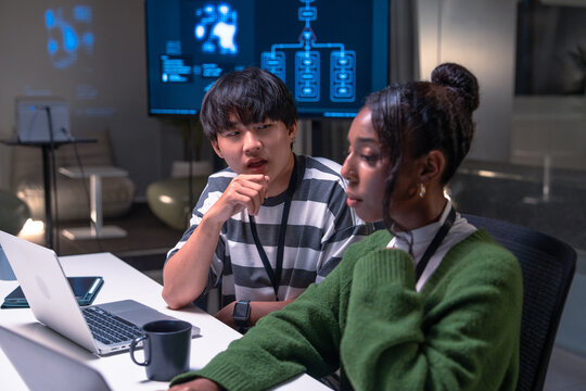Two programmer developers collaborate in office environment focusing on code development with laptops and digital screens, showing concentration and teamwork in modern workspace