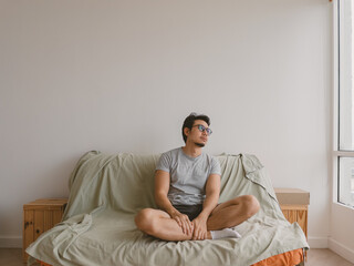 Asian man sitting on bed with legs crossed in minimalist bedroom in white shirt and clean background