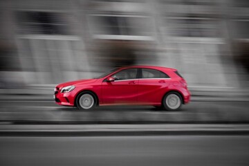 Obraz premium A sleek red hatchback races through a city street in motion blur, highlighting speed, style, and modern urban mobility against a muted background.