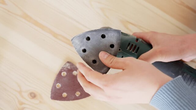 Hand changes sanding pad triangle on electric multitool placed over light wooden flooring