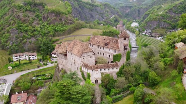 Aerial slow glide around Castel Roncolo (Schloss Runkelstein) in Bolzano, Italy, nestled above the Talvera river and surrounded by terraced vineyards and Alpine slopes