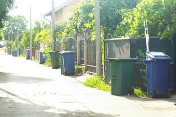 Green and blue plastic waste bins lined up along the fences of small private houses, illuminated by...