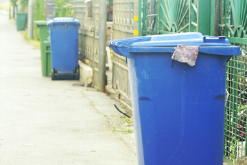 Fototapeta premium Green and blue waste bins on street lined up by families at the fences of their houses for garbage removal. Recycling culture, domestic trash segregation, eco responsibility and urban infrastructure