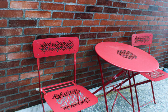 Red metal outdoor table and two chairs with decorative patterns are positioned in front of a rustic red brick wall - Powered by Adobe