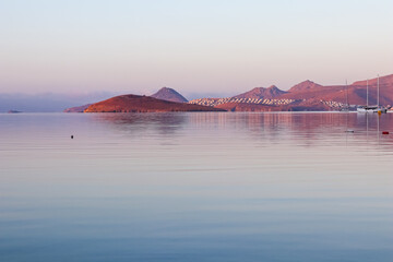 A pastel-hued sunrise or sunset over a tranquil sea, with a rocky island and distant coastal towns...