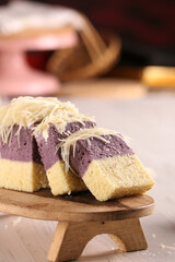 A sliced two-layer cake, featuring a taro top layer and a lighter bottom layer, is generously topped with grated cheese and presented on a small wooden stand