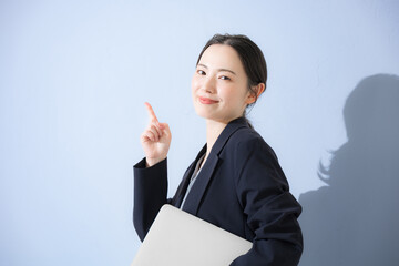 Pointing to a standing point etc. wearing a suit with a computer, young cute woman's business upper body Light blue background Sales and other recruiting images