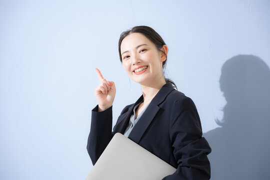パソコン持ってスーツを着た立つポイントなどの指差しポーズの若いかわいい女性のビジネス上半身　水色背景の営業などリクルートのイメージ