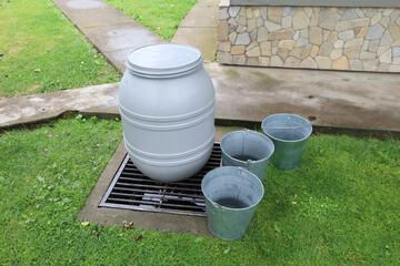 A gray plastic barrel for collecting rainwater stands on metal grates, with three galvanized...