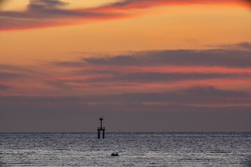 A striking seascape capturing the vivid hues of a sunset sky blending orange, red, and purple over the calm ocean. A navigation tower stands against the horizon, adding depth and dramatic scene.