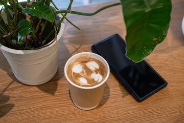 Close-up of Hot coffee latte with latte art milk foam in cup made of paper and smartphone on wood...
