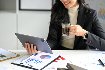 Businesswoman reviewing charts and reports on digital tablet while enjoying a cup of coffee at desk