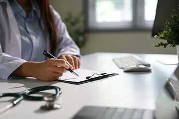 Female doctor writing on a clipboard at her desk during a consultation in a clinical office