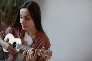 Woman sitting indoors playing the ukulele with a calm and focused look, dressed casually, enjoying...