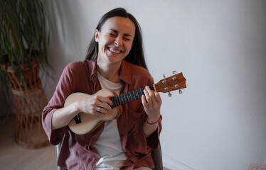 Cheerful young woman laughing while playing ukulele at home, eyes closed in joy, wearing casual...