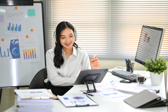 Asian employee wearing headphones attends an online meeting using digital tablet at desk