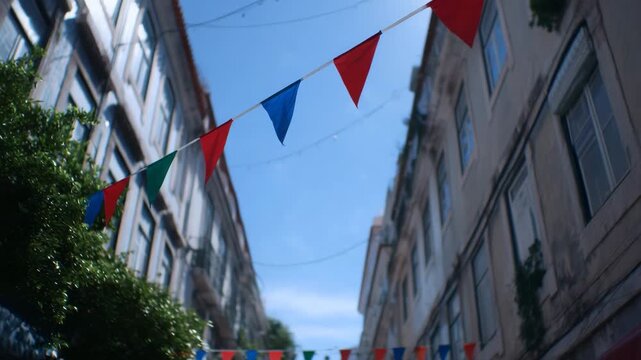 Festive street adorned with colorful flags against a bright blue sky. The flags are strung above a street lined with buildings, creating a celebratory atmosphere