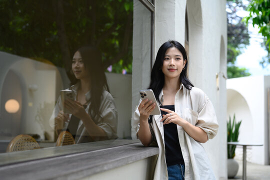 Stylish young woman standing outside a cafe while holding her smartphone