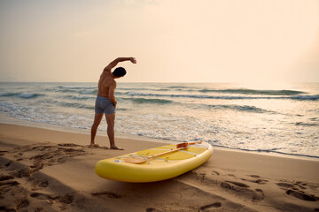 Man stretching on beach beside paddleboard at sunrise
