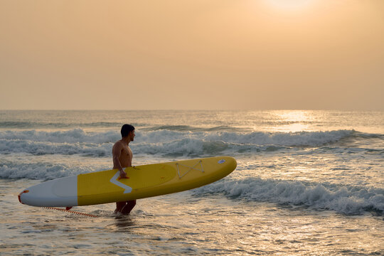 Man carrying paddleboard at sunrise on beach