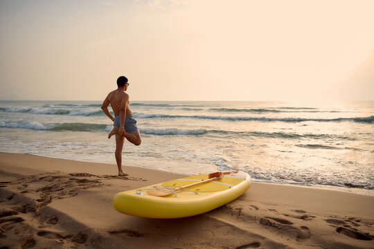 Surfer stretching on the beach at sunset with paddle board
