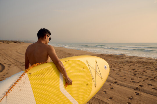 Surfer carrying paddle board walking on the beach at sunset